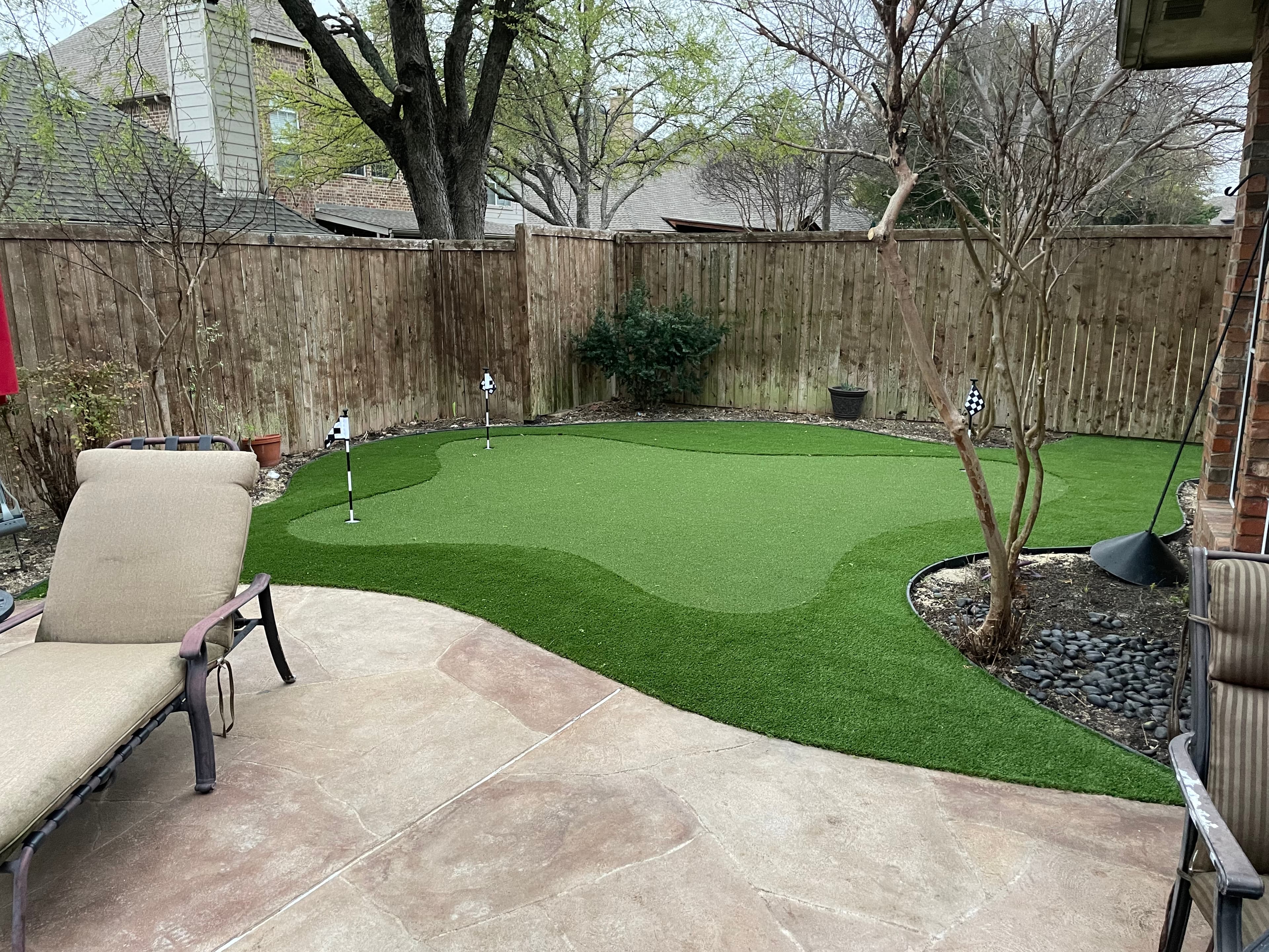Backyard artificial turf putting green with three holes, a stone patio, and a lounge chair.