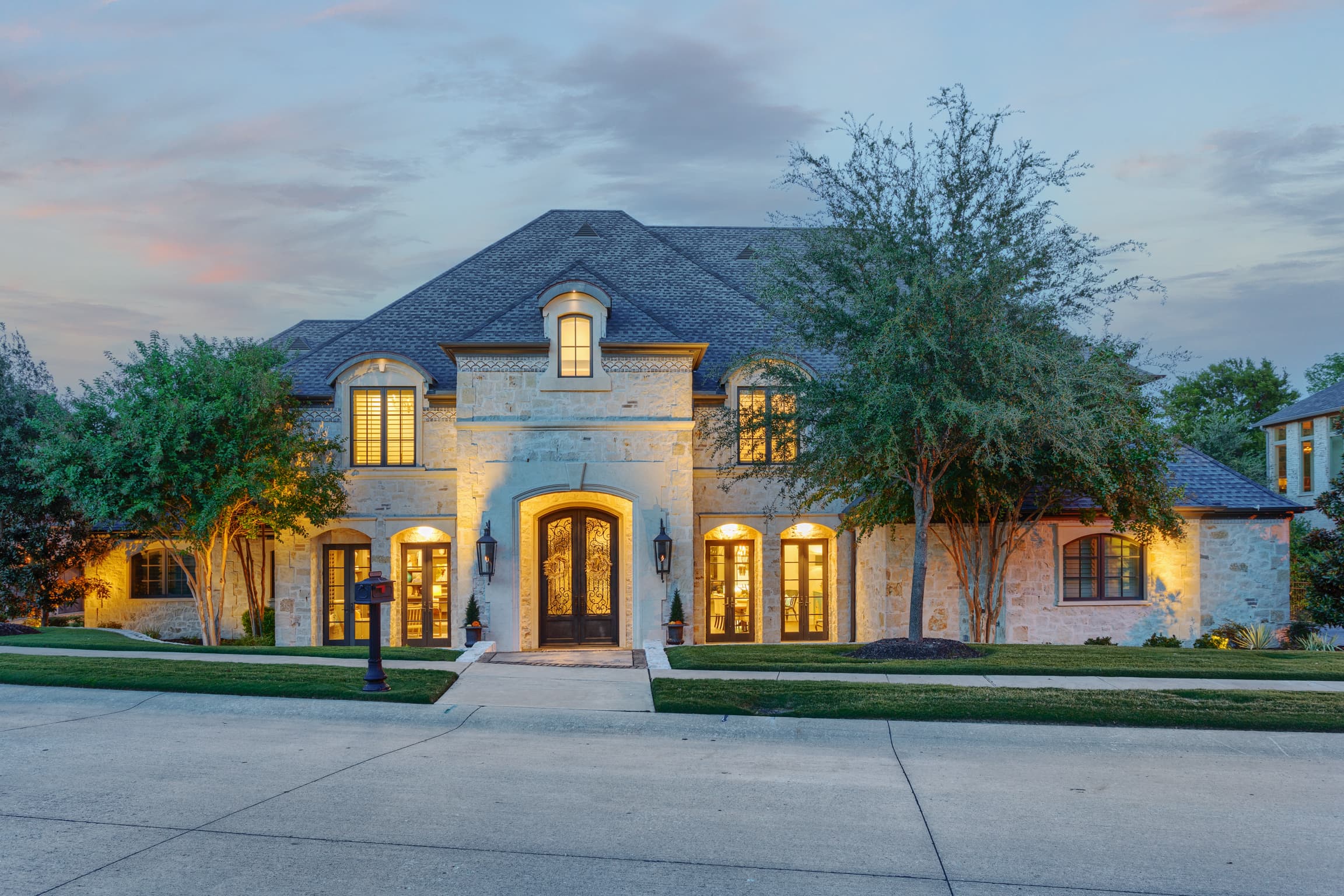 Luxurious stone mansion at dusk with warm interior lights glowing through large windows and doors.