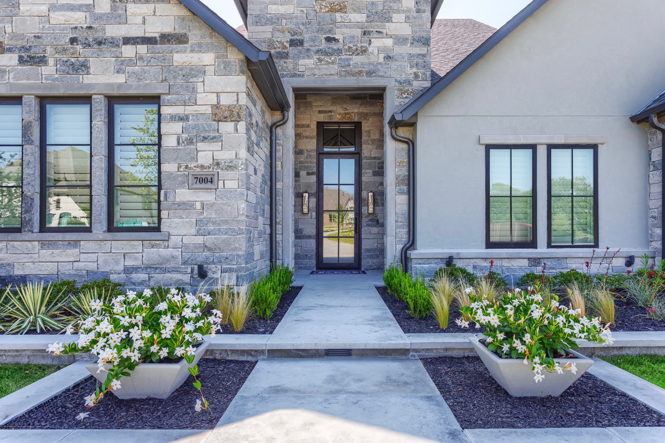 Modern house entrance with grey stone walls, glass door, and white flowers in concrete planters.