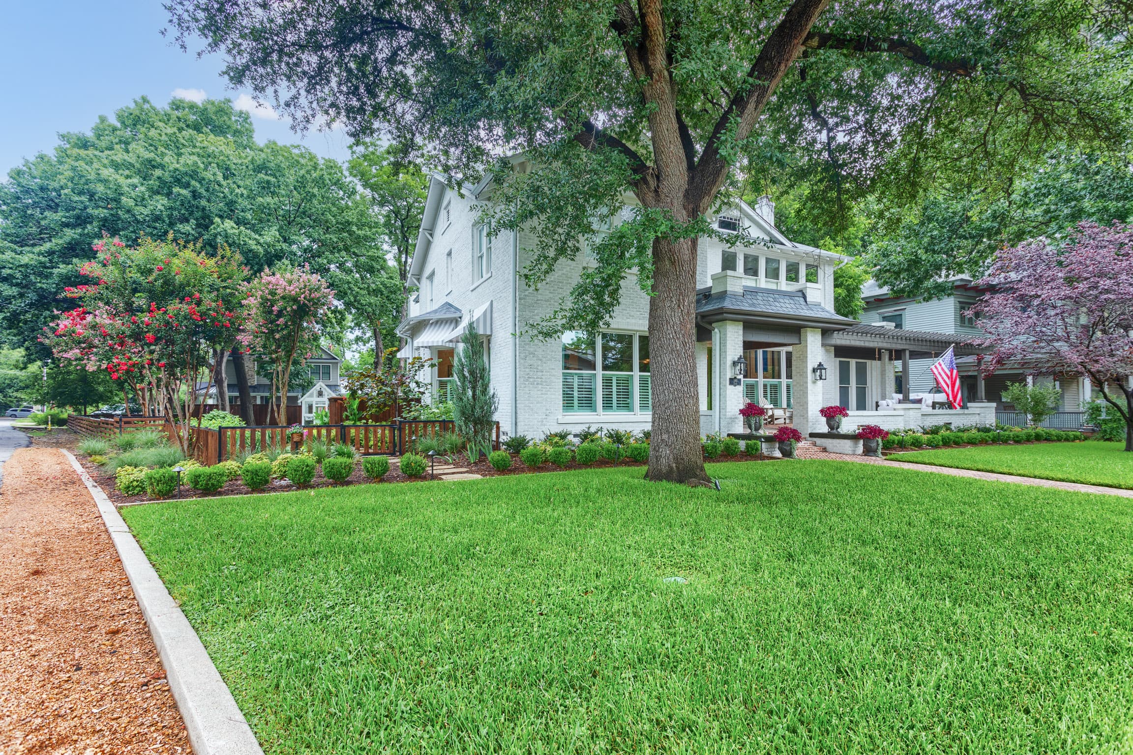 White brick house with a sprawling green lawn, large oak tree, and colorful landscaping.