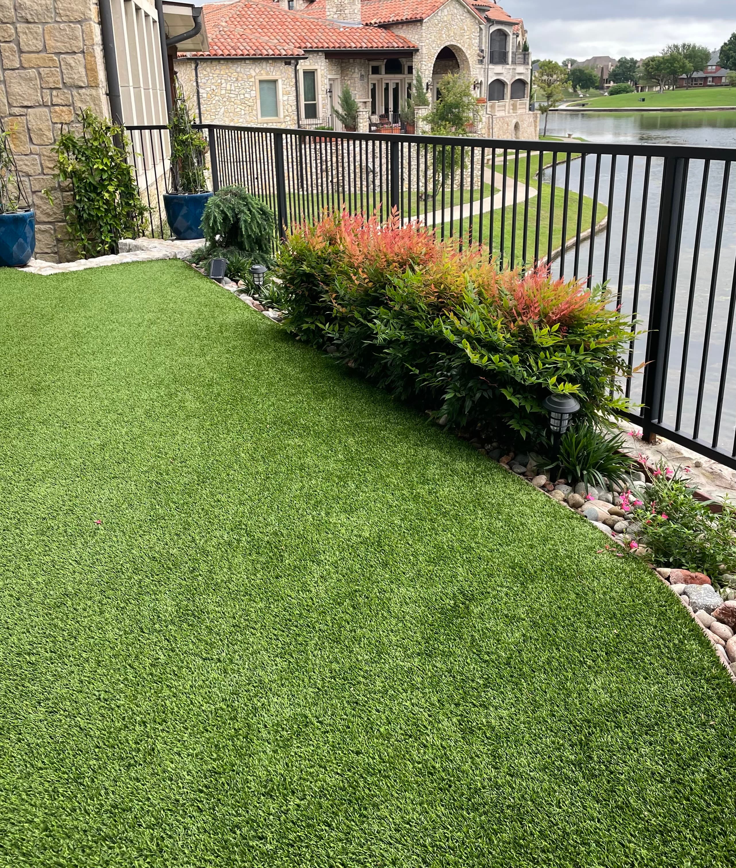Lush artificial turf lawn and bushes along a black fence overlooking a scenic lakefront.