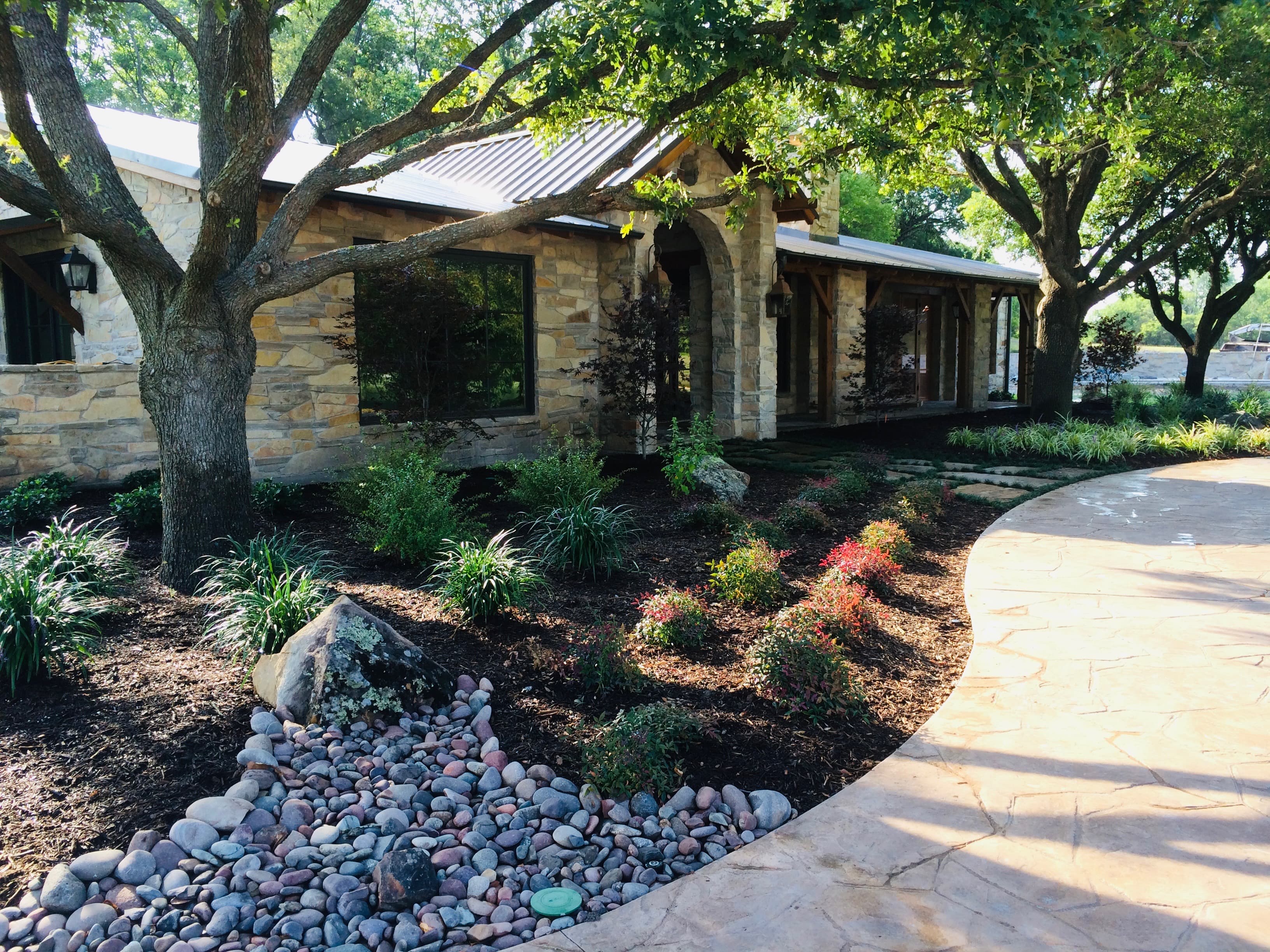 Stone house with metal roof and arched entryway, featuring a curved walkway and lush landscaping.