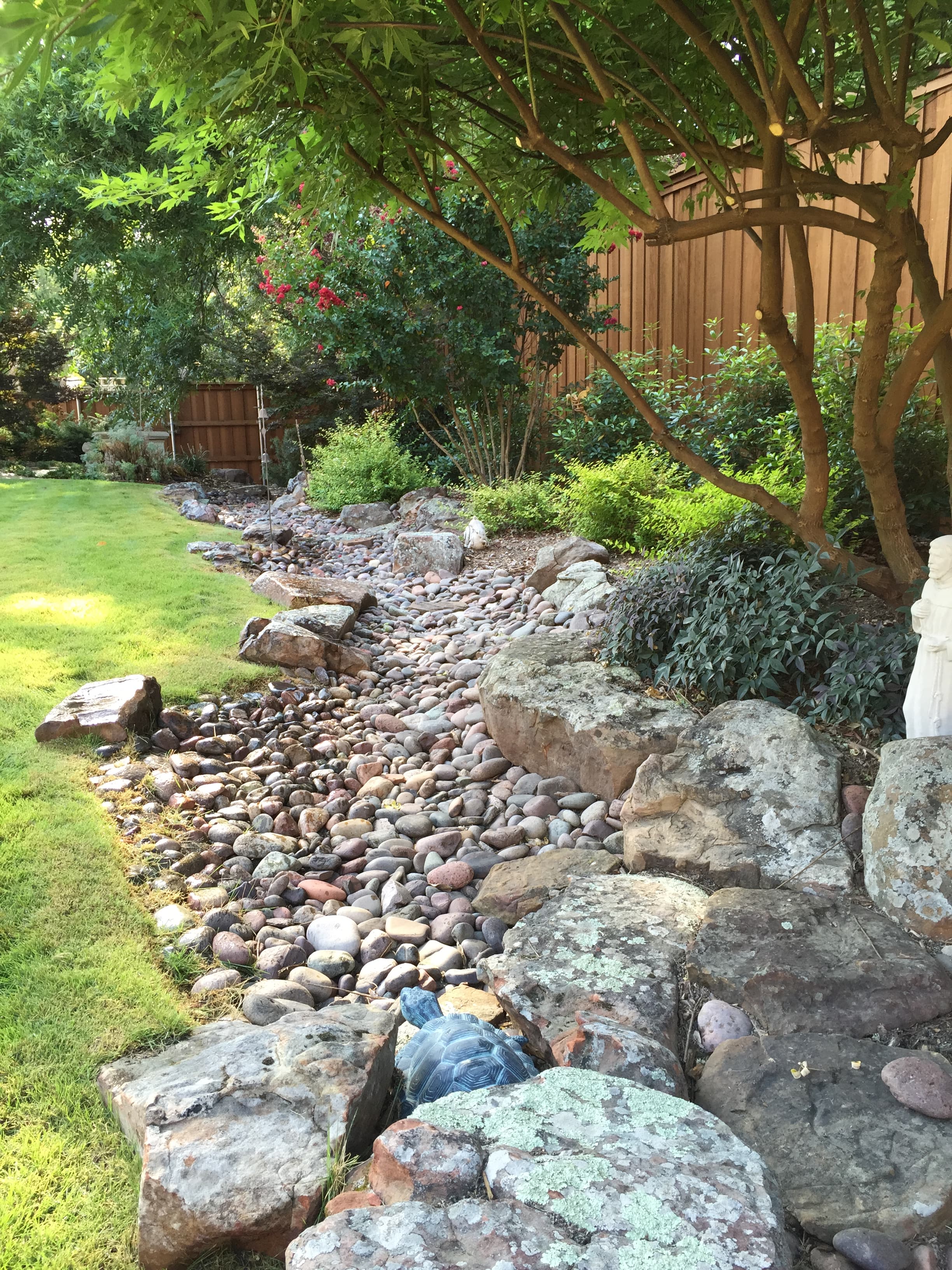 Dry creek bed of smooth river stones winding through a lush, green landscaped garden.