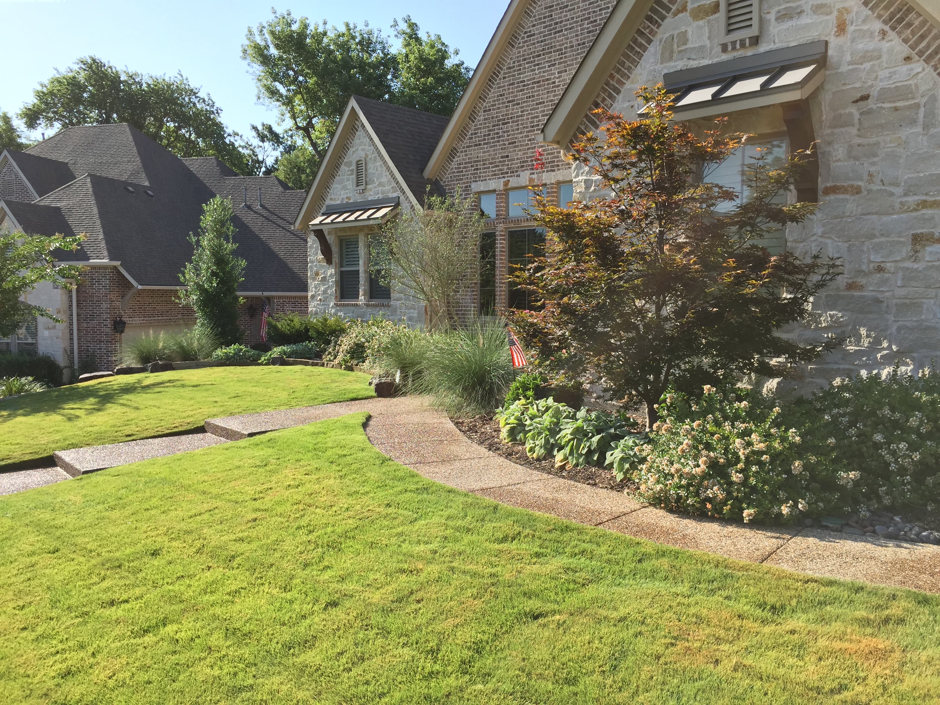 Suburban stone house featuring a manicured lawn, curved walkway, and lush garden landscaping.