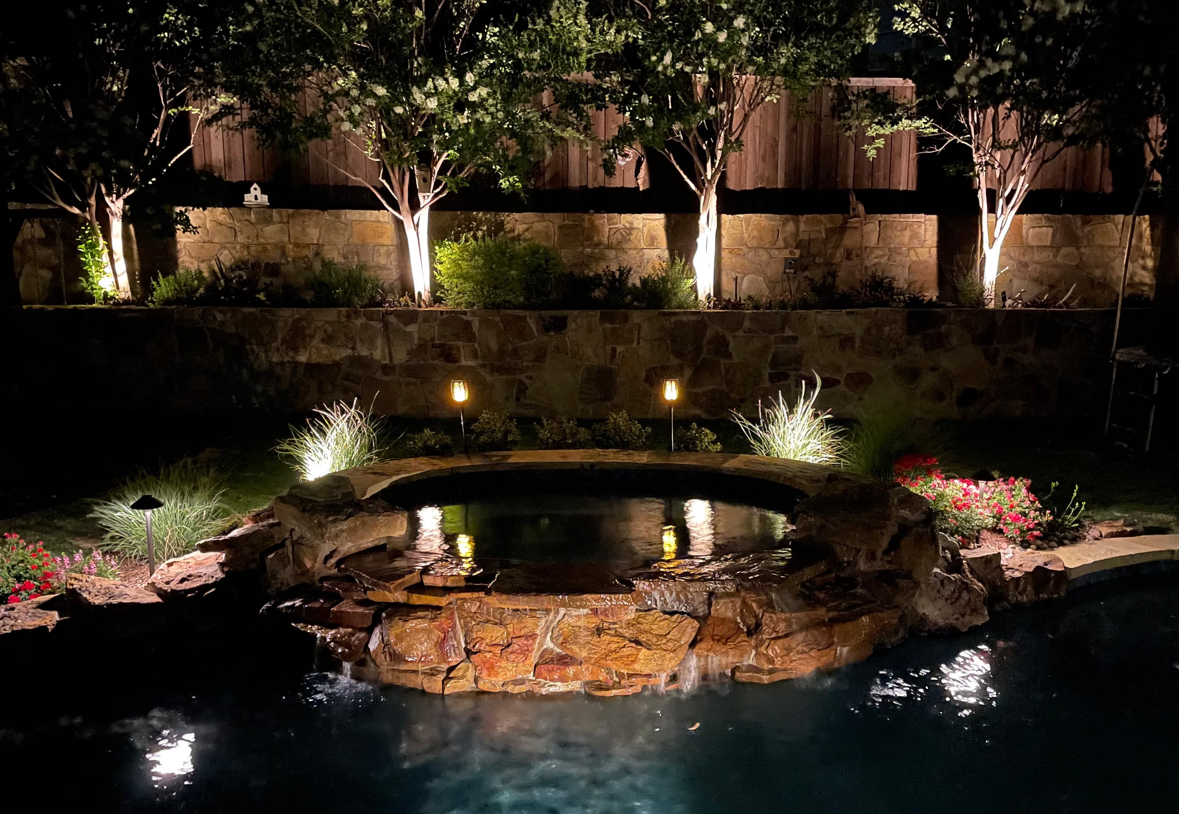 Illuminated stone hot tub with waterfall into a swimming pool in a nighttime backyard.