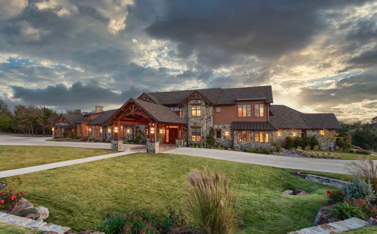 Sprawling rustic stone mansion with warm glowing windows under a dramatic cloudy sky at sunset.