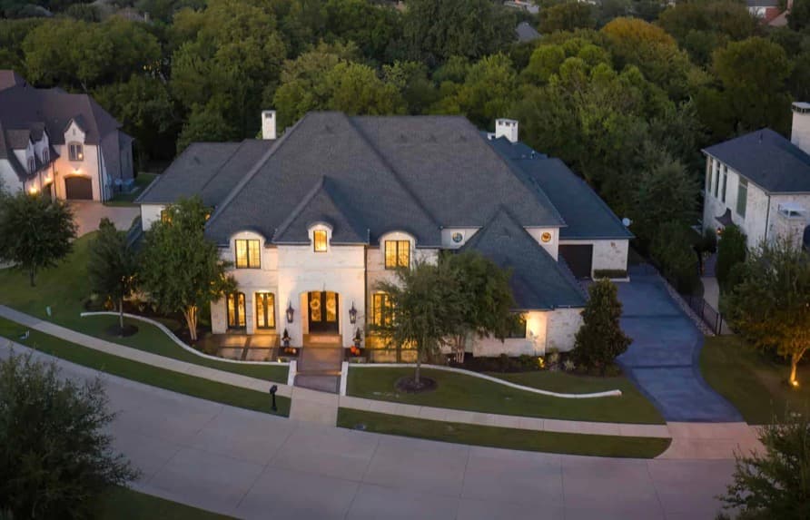 Aerial view of a luxury white stone home with lit windows at dusk among trees.