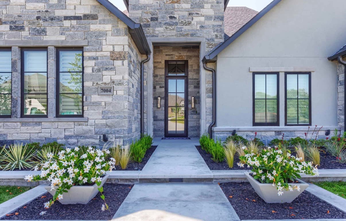 Modern house entrance featuring stone walls, glass door, concrete walkway, and white flowers in planters.