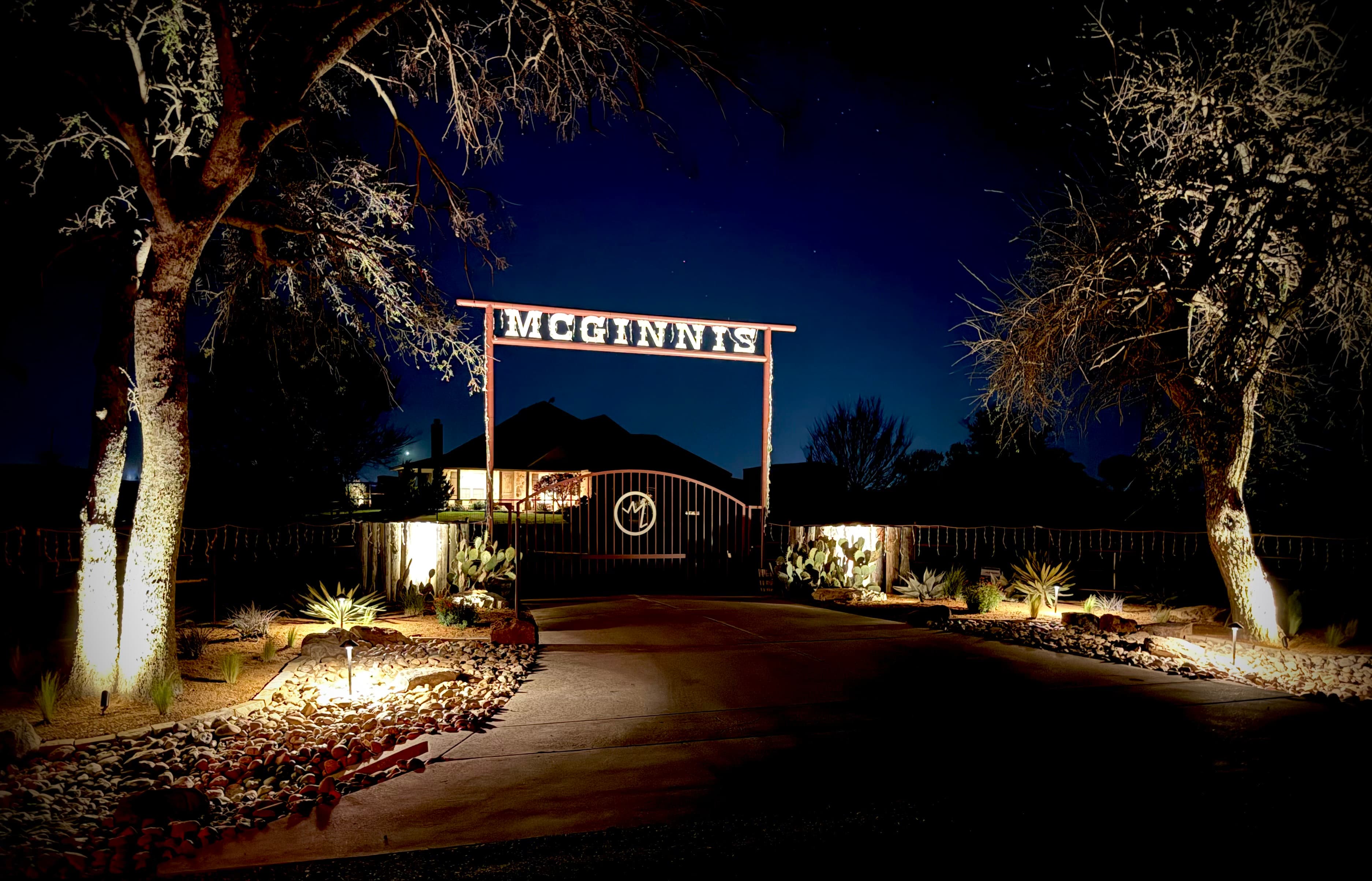 Illuminated McGinnis ranch gate at night with desert landscaping and trees lit from below.