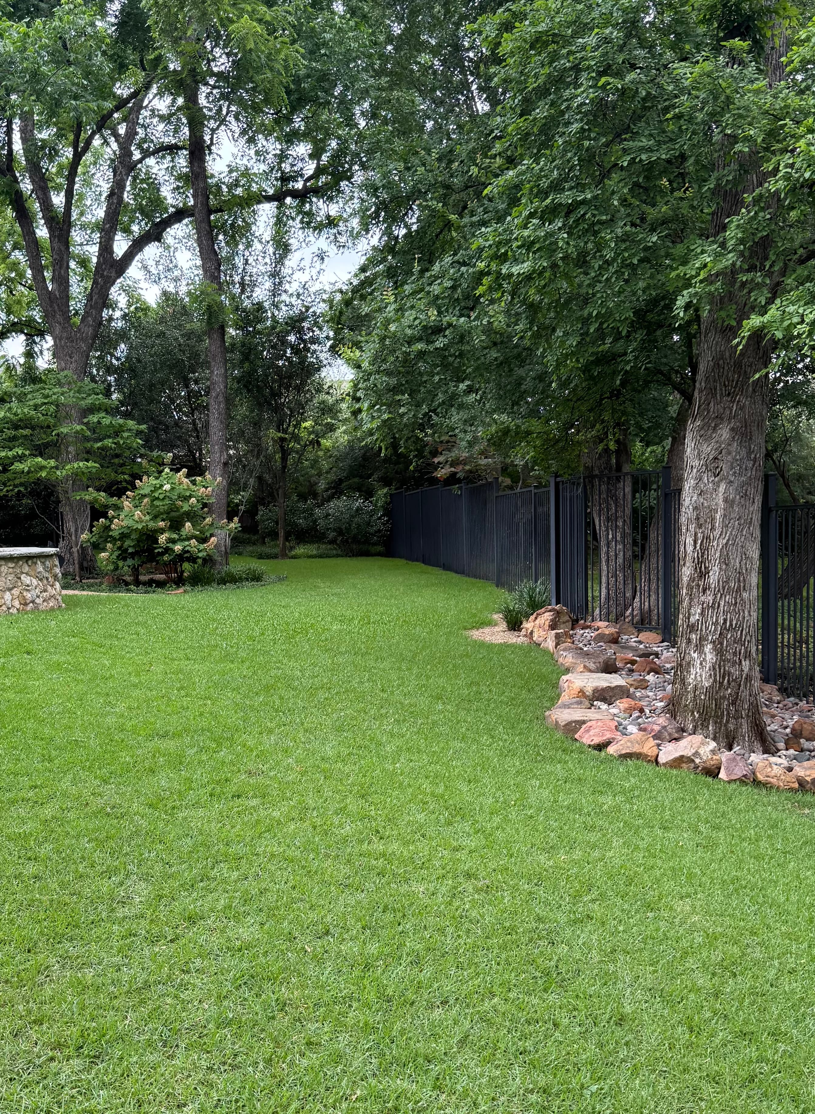 Lush green lawn bordered by a black metal fence and mature trees with stone edging.