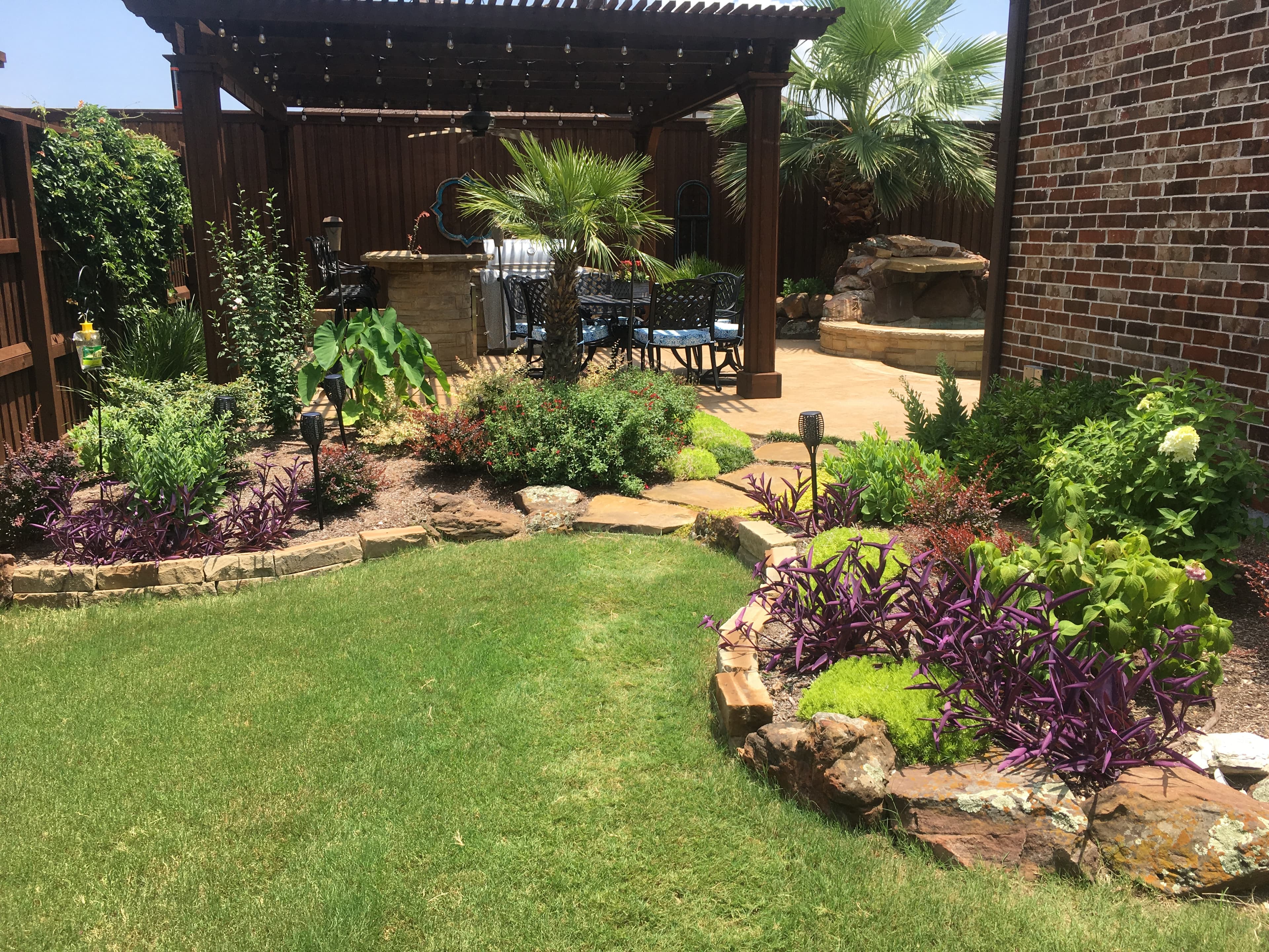 Landscaped backyard with a green lawn, purple plants, and a wooden pergola over a patio.