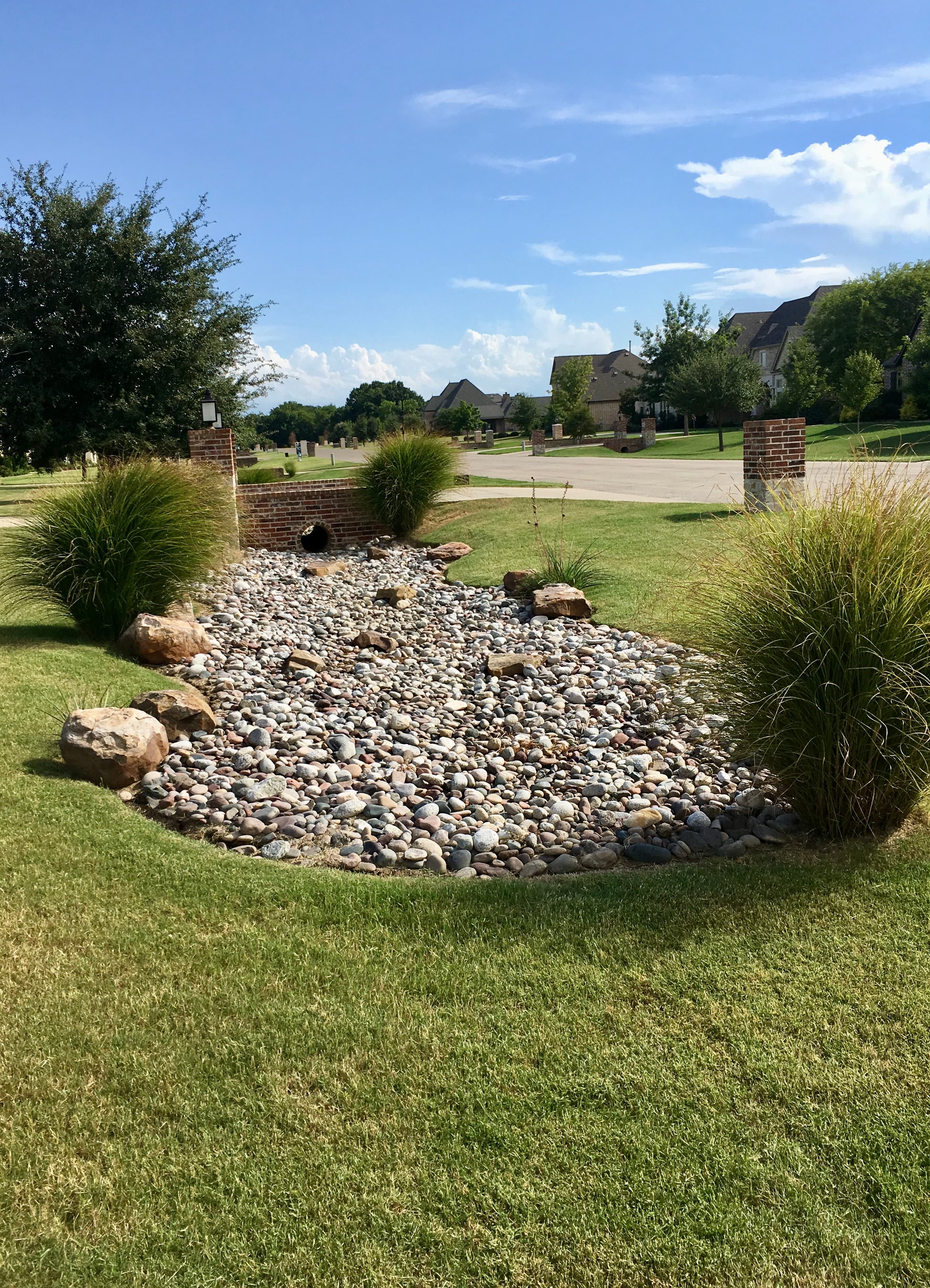 Landscaped dry creek bed with river rocks and ornamental grasses in a suburban neighborhood.