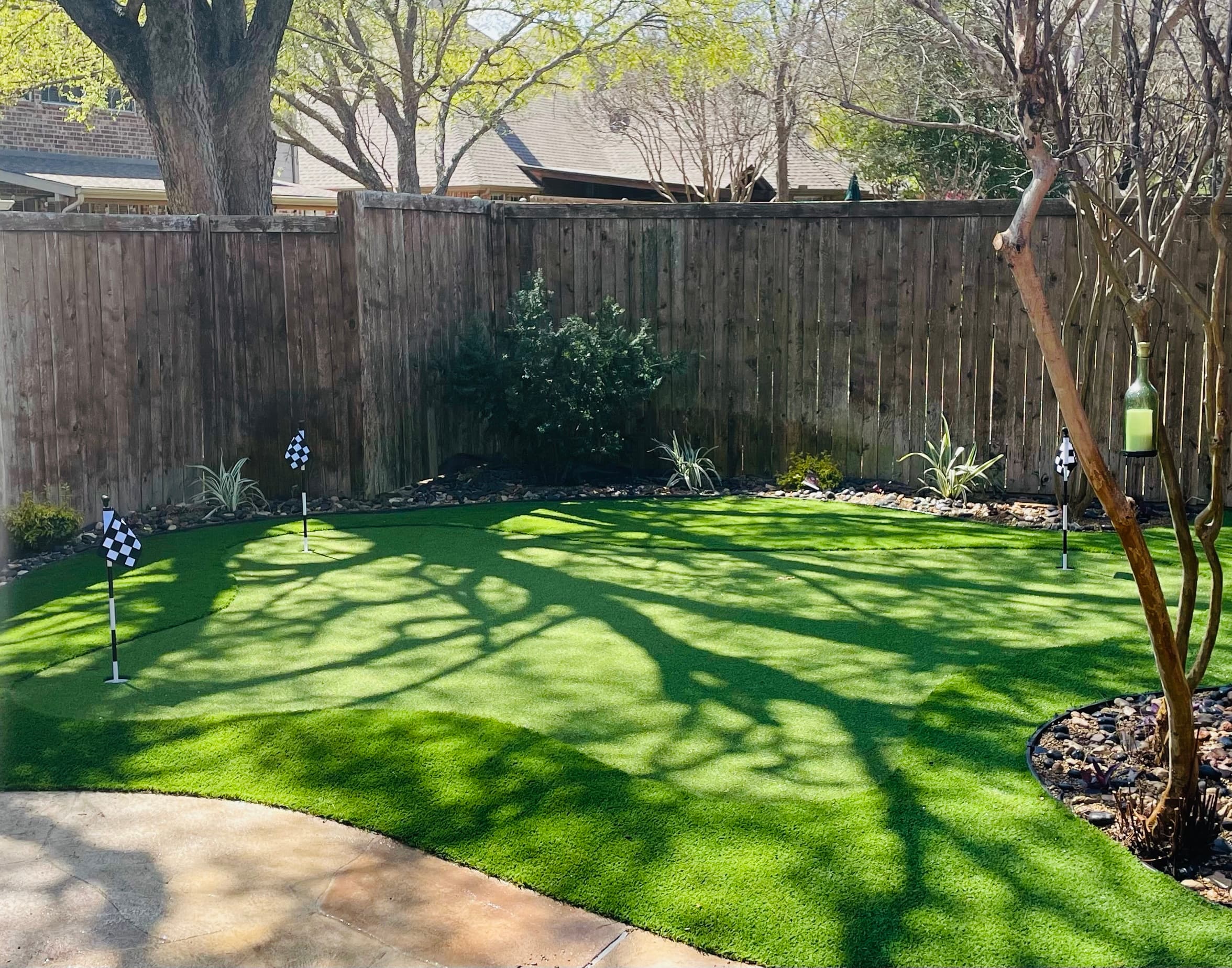 Backyard putting green with artificial turf, three checkered flags, and shadows from surrounding trees.