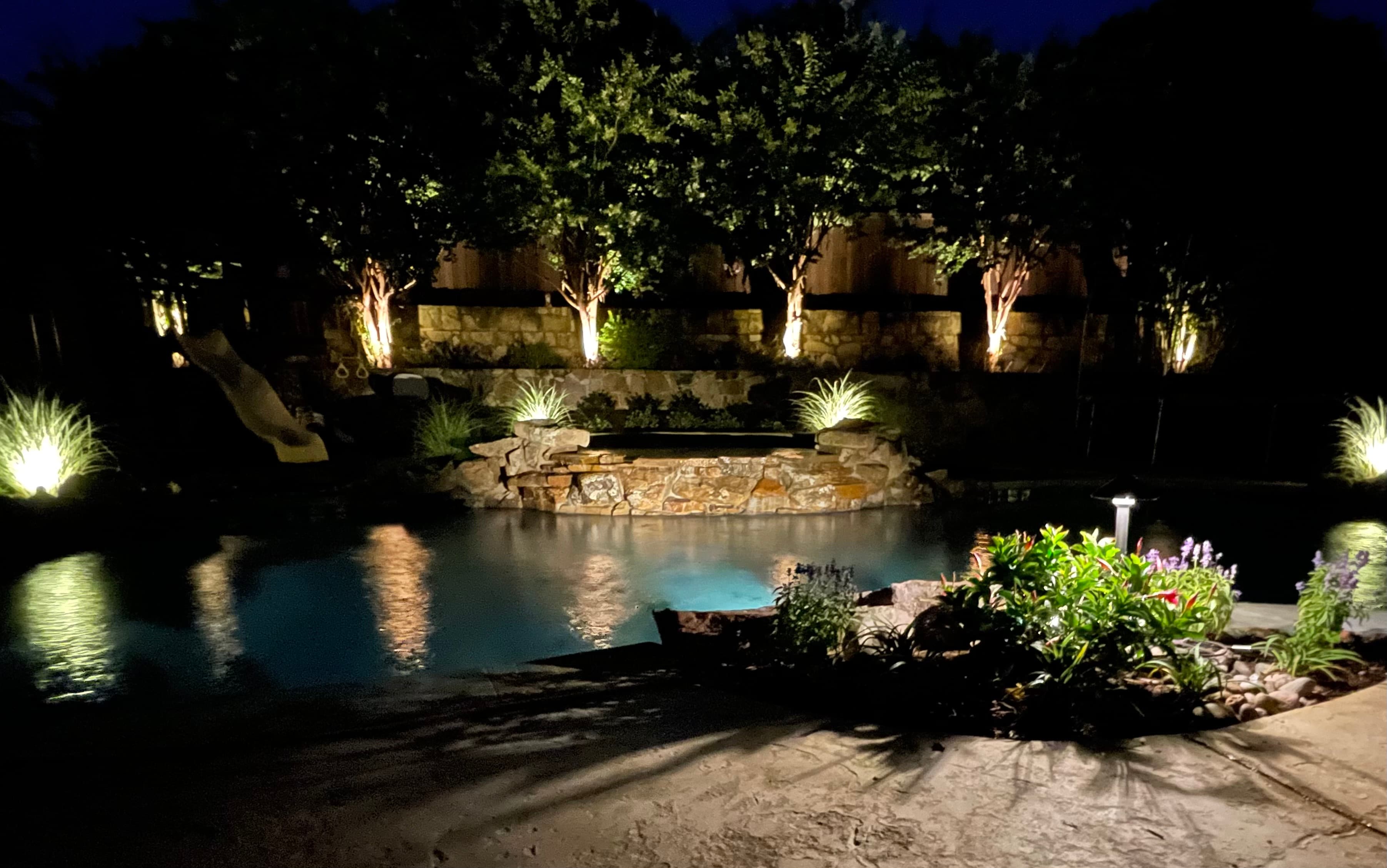 Backyard pool at night with a stone waterfall, glowing landscape lights, and illuminated trees.