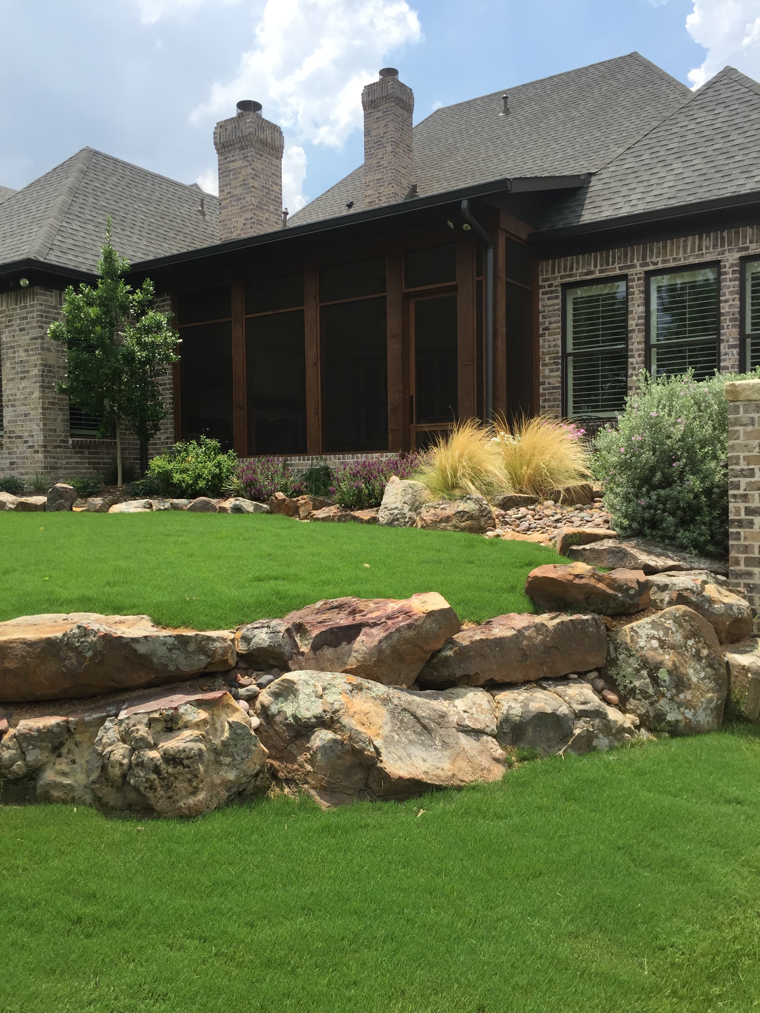 Brick house with a screened porch, lush lawn, and a large natural stone retaining wall.