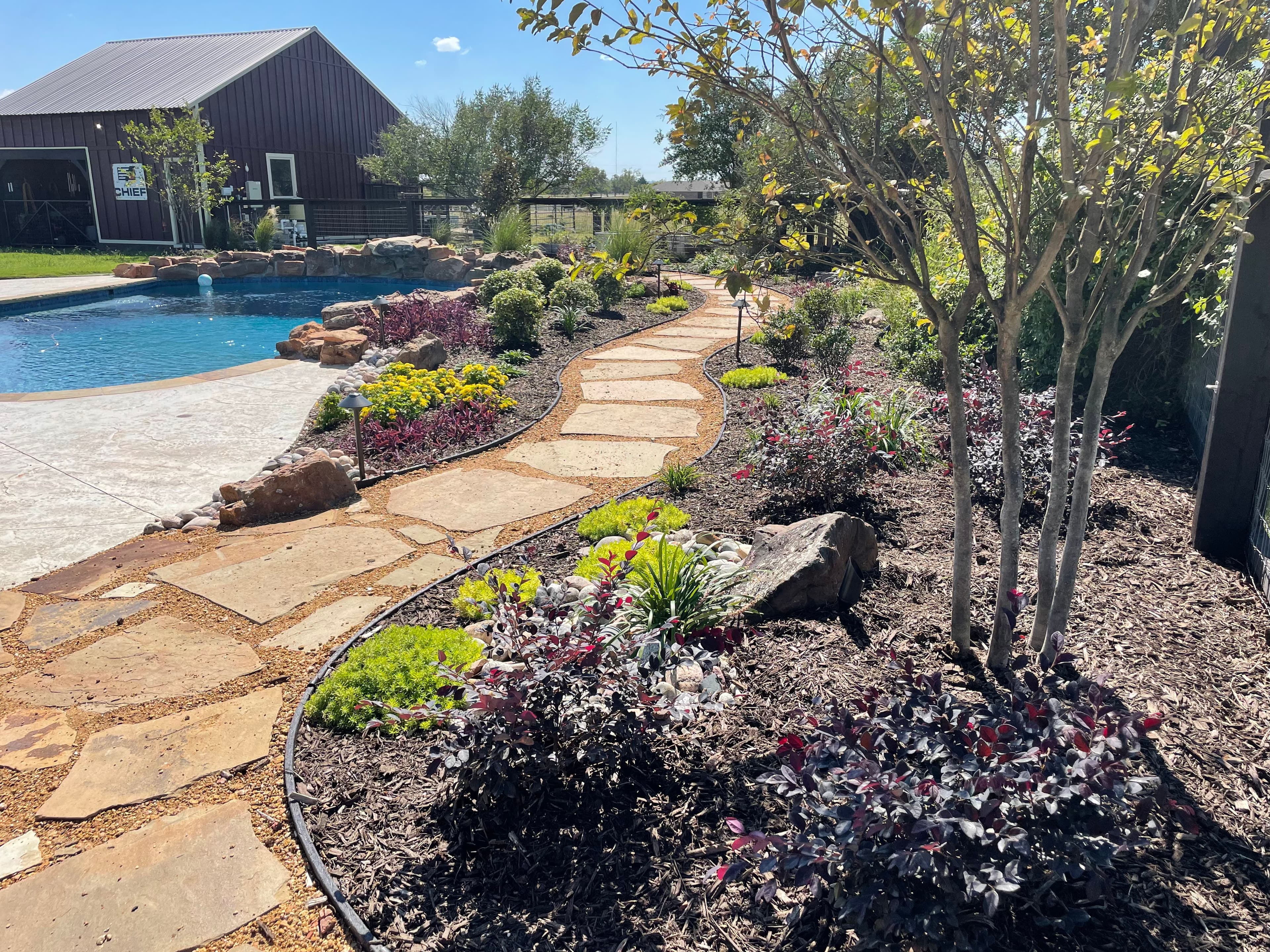 Curved flagstone path through lush garden beds beside a swimming pool and large barn.