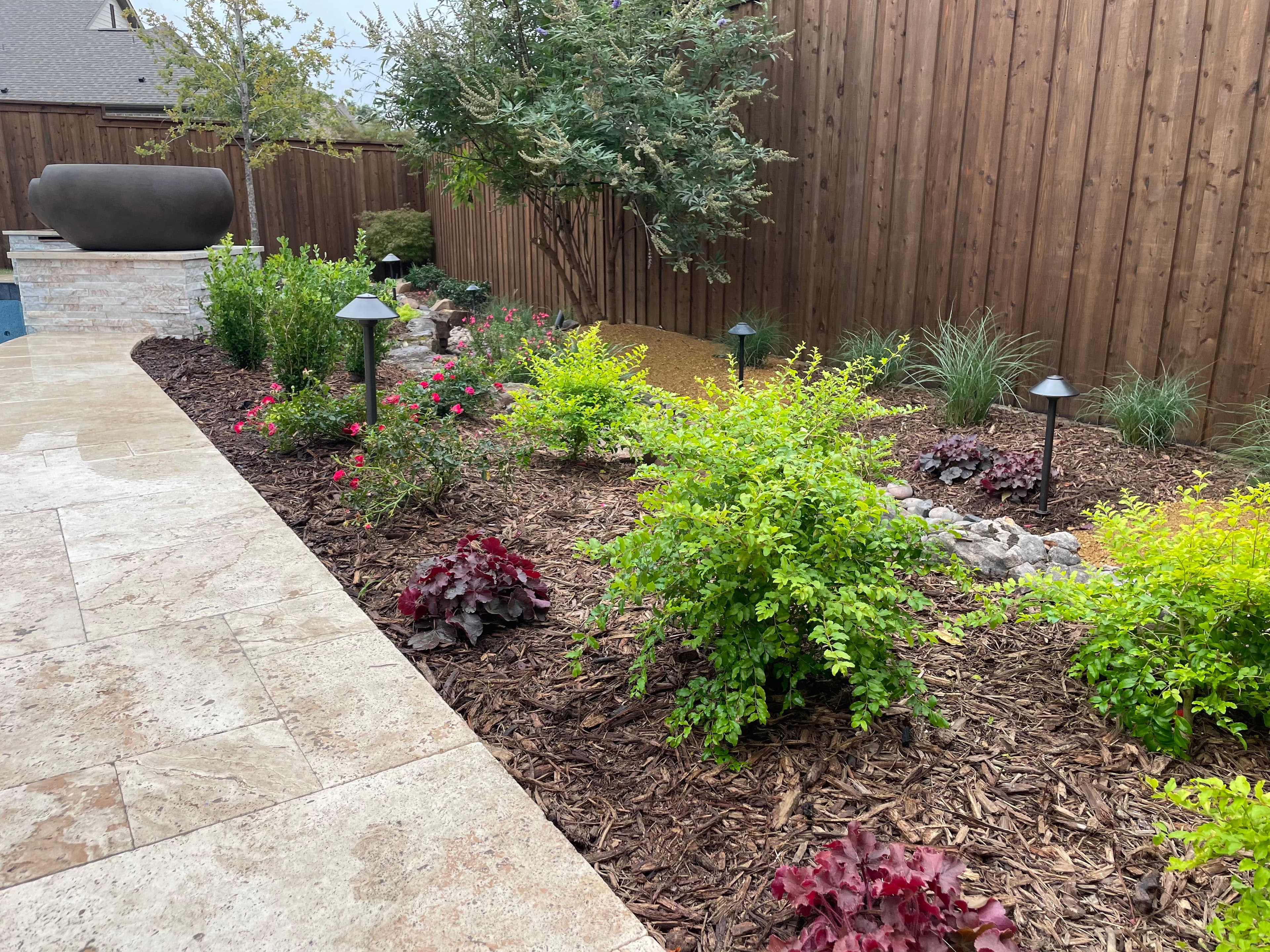 Lush garden bed with green shrubs and path lights bordering a light stone tile walkway.