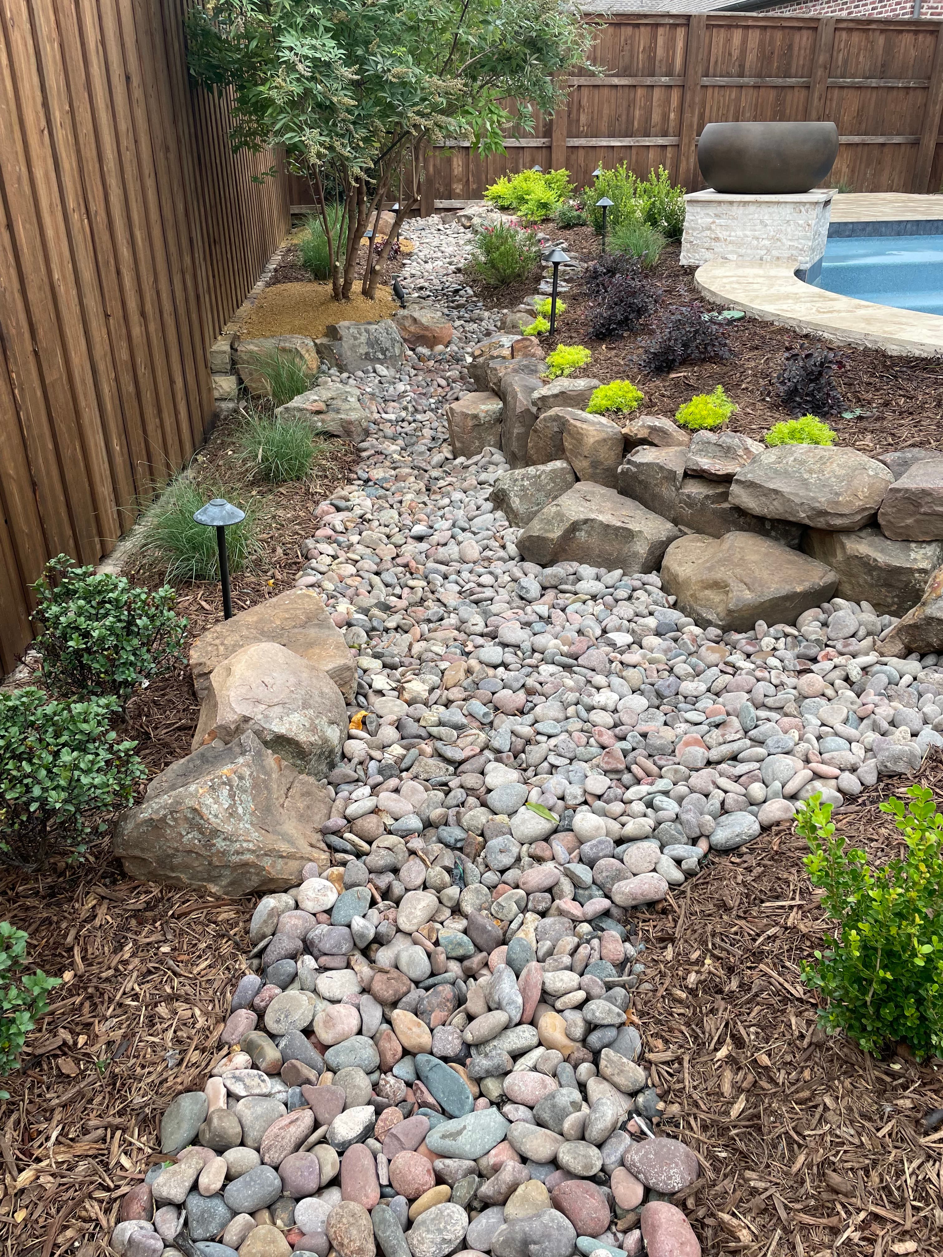 Landscaped garden featuring a dry creek bed of river rocks, boulders, mulch, and lush greenery.