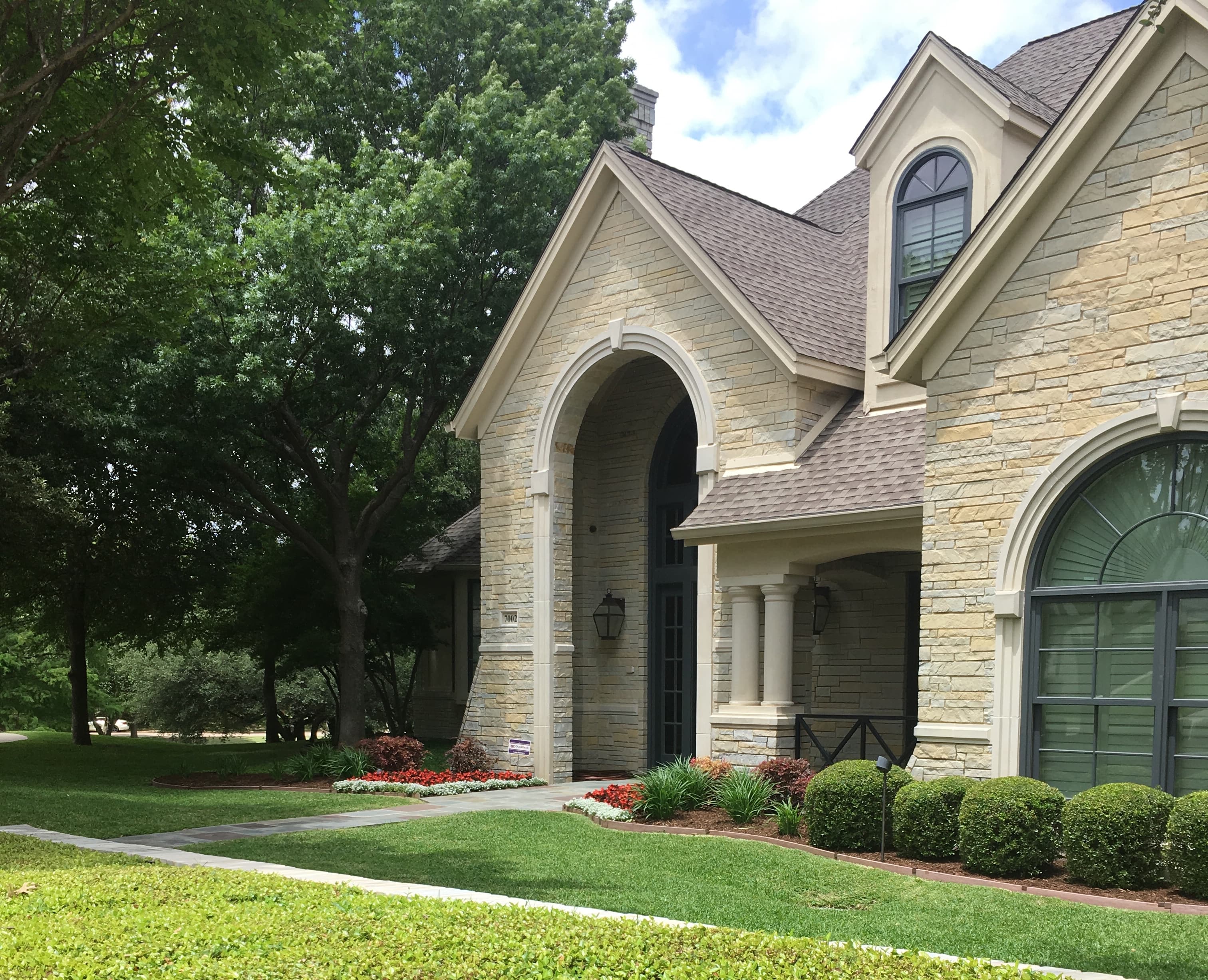 Luxurious light stone house with arched windows and entryway, surrounded by lush green landscaping.