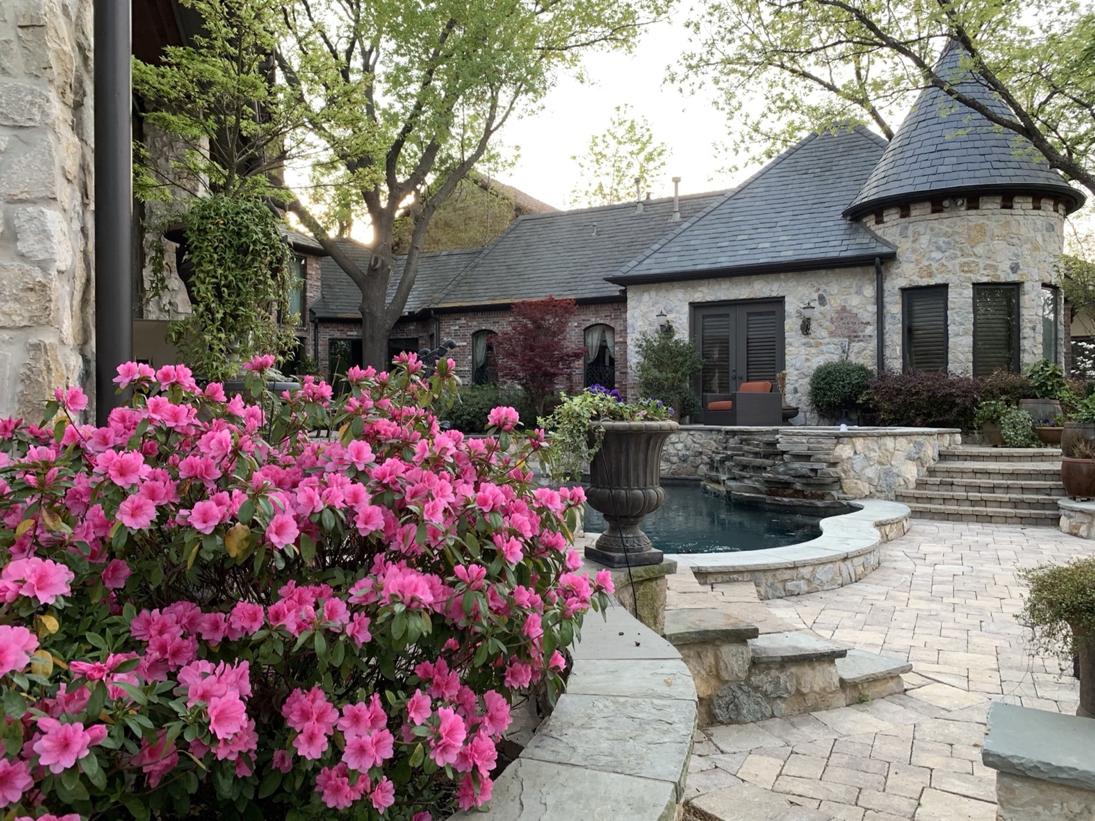 Vibrant pink azaleas bloom in a stone-paved backyard with a pool and turreted house.